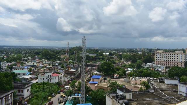 A panoramic view of a cityscape featuring buildings, trees, and telecommunications towers under a cloudy sky.