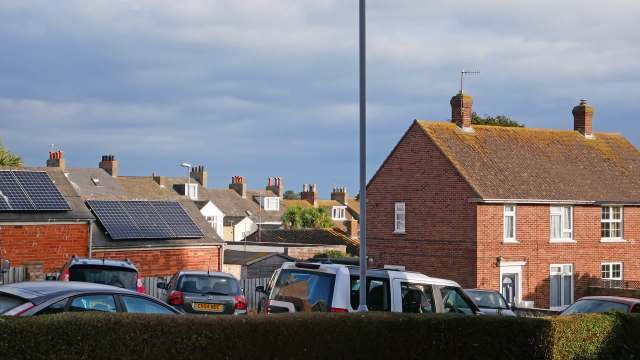 Houses with solar panels and parked cars in an urban area under a partly cloudy sky.