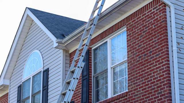 A ladder leans against the side of a two-story brick and siding house with multiple windows.