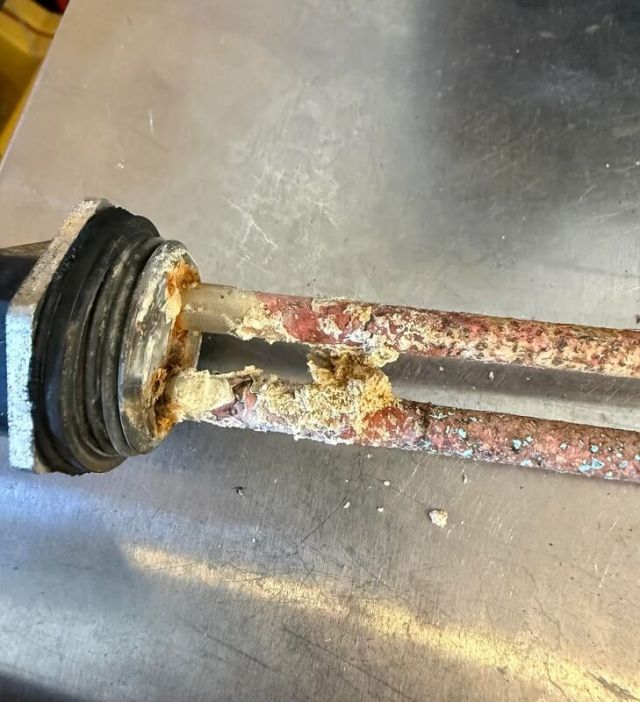 A close-up of a corroded heating element attached to a black circular base, positioned near metallic fittings.