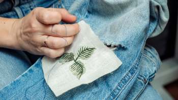 A hand stitches a fabric patch with an embroidered leaf design onto a pair of blue denim jeans.