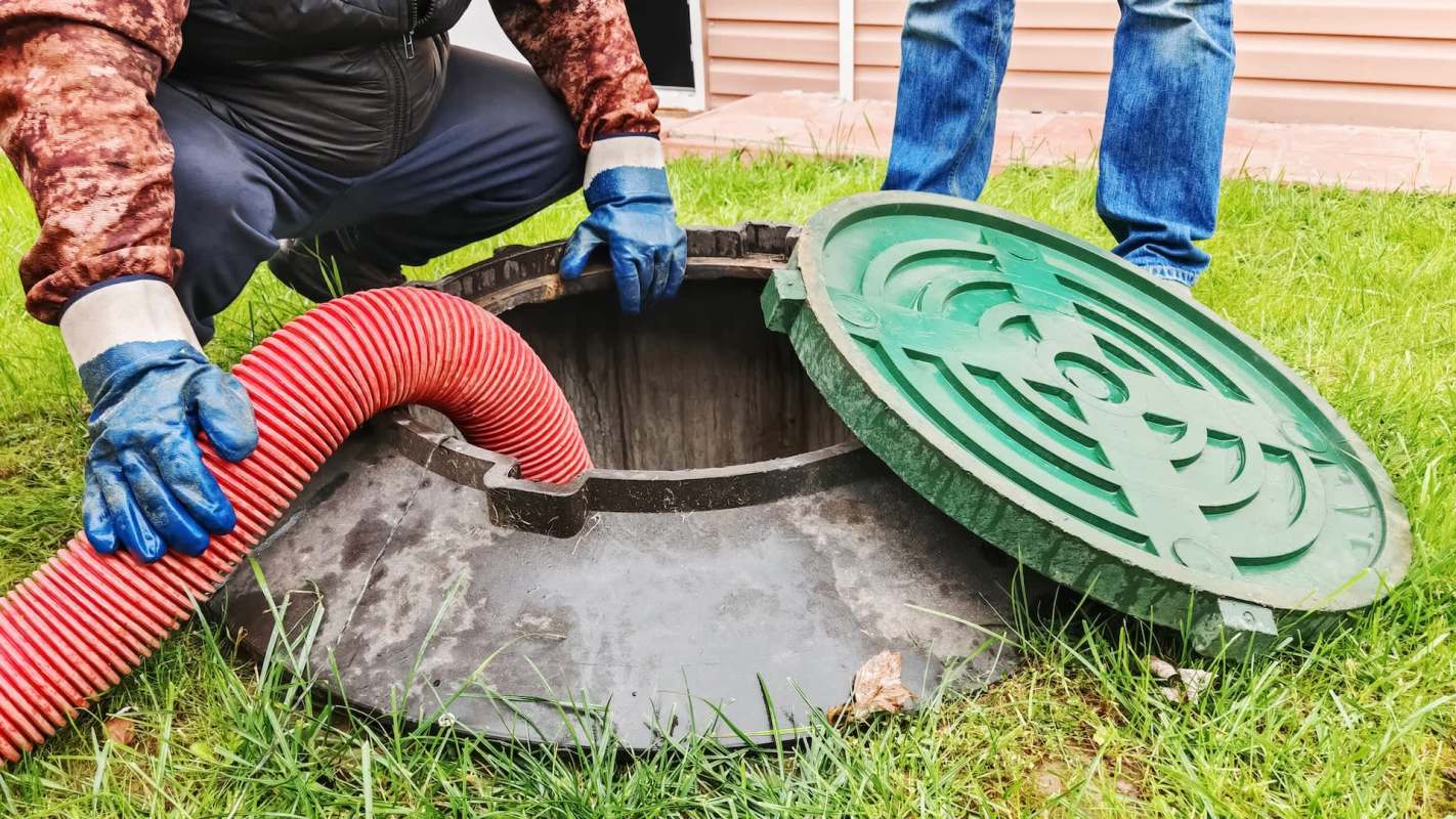 Two people using a red hose to access a manhole cover in a grassy area.