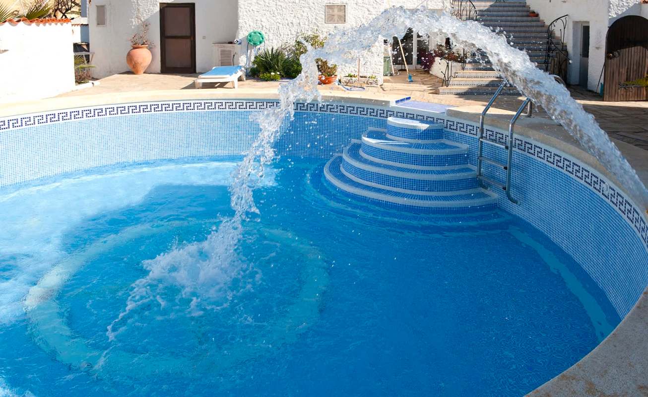 A water feature sprays into a blue swimming pool with steps and a nearby house in the background.
