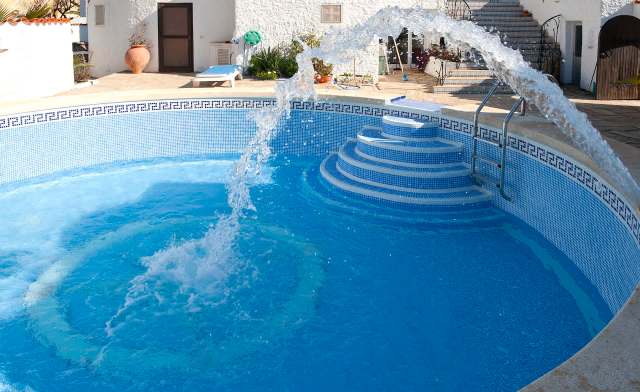 A water feature sprays into a blue swimming pool with steps and a nearby house in the background.