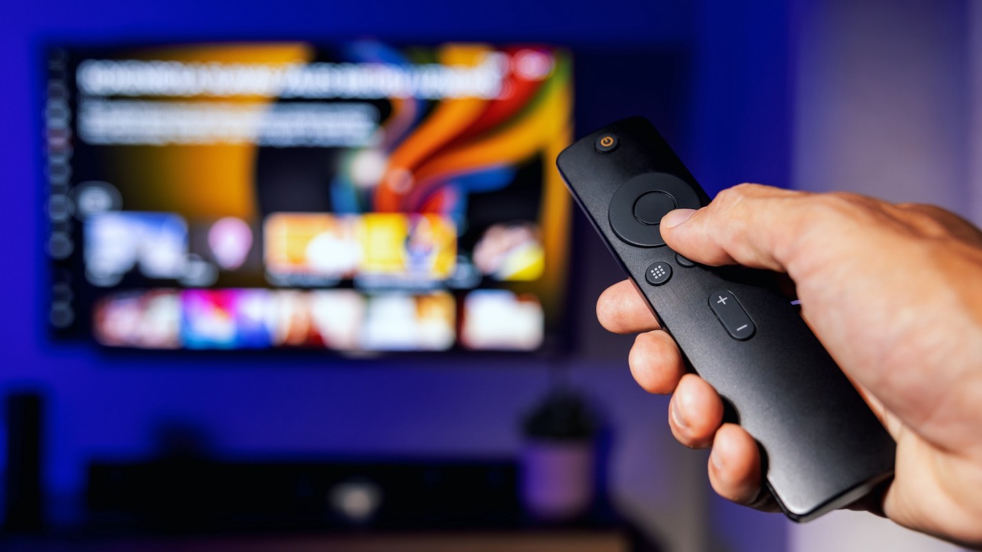 A hand holds a remote control in front of a blurred television screen displaying various content options.