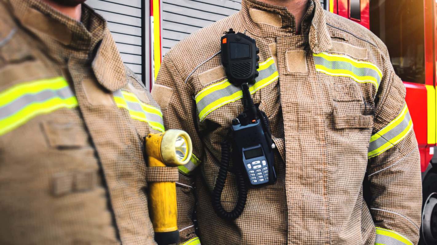 Two firefighters in uniforms with reflective stripes, standing near a fire truck, equipped with a flashlight and radio.