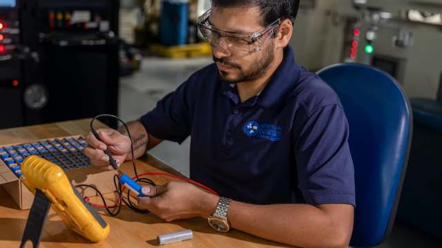 A man tests a battery using a multimeter at a workbench.