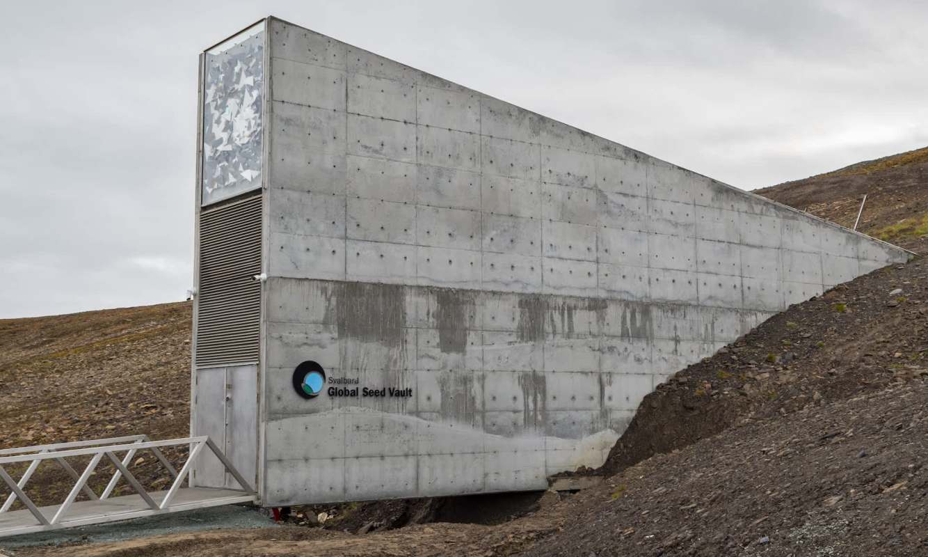 A concrete building with a sloped roof and a sign reading "Svalbard Global Seed Vault" on a rocky landscape.