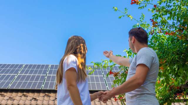 A man and a woman stand together, holding hands and looking at solar panels on a roof under a clear blue sky.