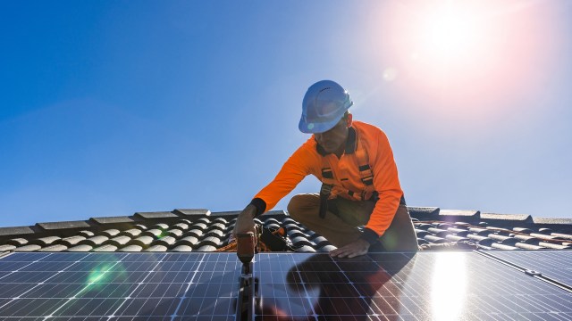 A worker installs solar panels on a roof under a clear blue sky.