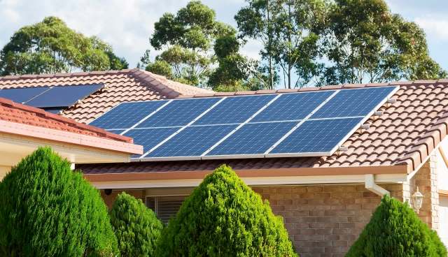 Solar panels installed on the roofs of two houses surrounded by hedges and trees.