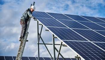 A worker standing on a ladder installs solar panels under a blue sky.