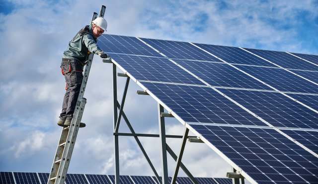 A worker standing on a ladder installs solar panels under a blue sky.