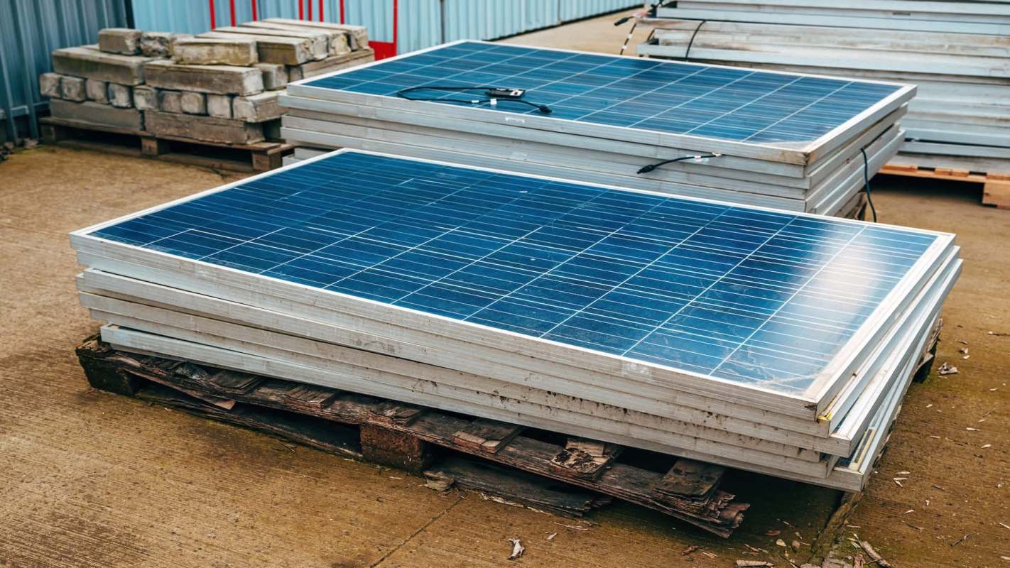 Stacked solar panels resting on pallets in an outdoor storage area.