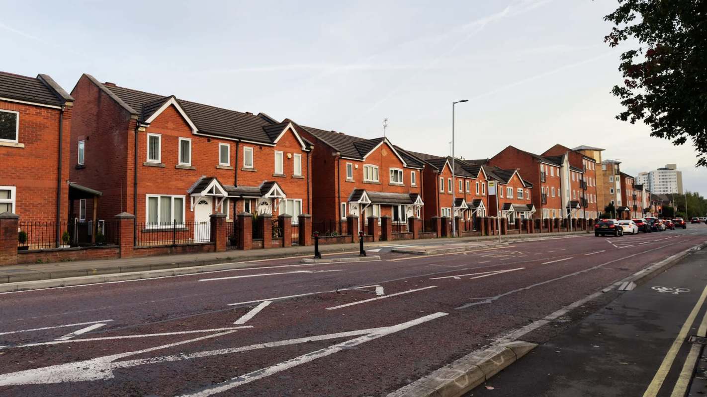 A row of red brick houses along a road with parked cars and pavement.