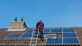 A worker on a ladder installs solar panels on a rooftop under a clear blue sky.
