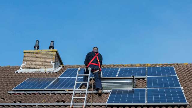 A worker on a ladder installs solar panels on a rooftop under a clear blue sky.