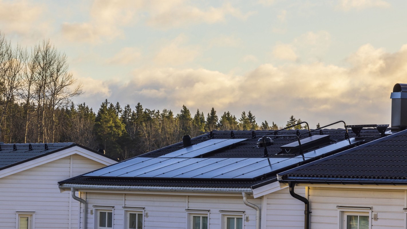 Roofs of white houses with solar panels and a backdrop of trees and cloudy skies.