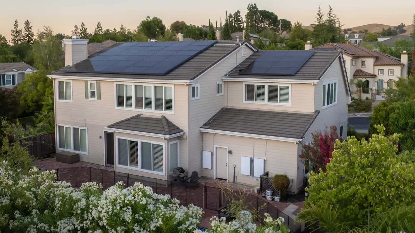 A two-story house with solar panels on the roof, surrounded by greenery and flowers.