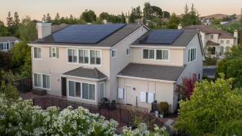 A two-story house with solar panels on the roof, surrounded by greenery and flowers.