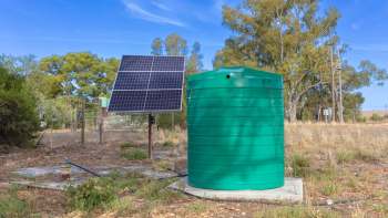 A green water tank next to a solar panel in a grassy area with sparse trees.