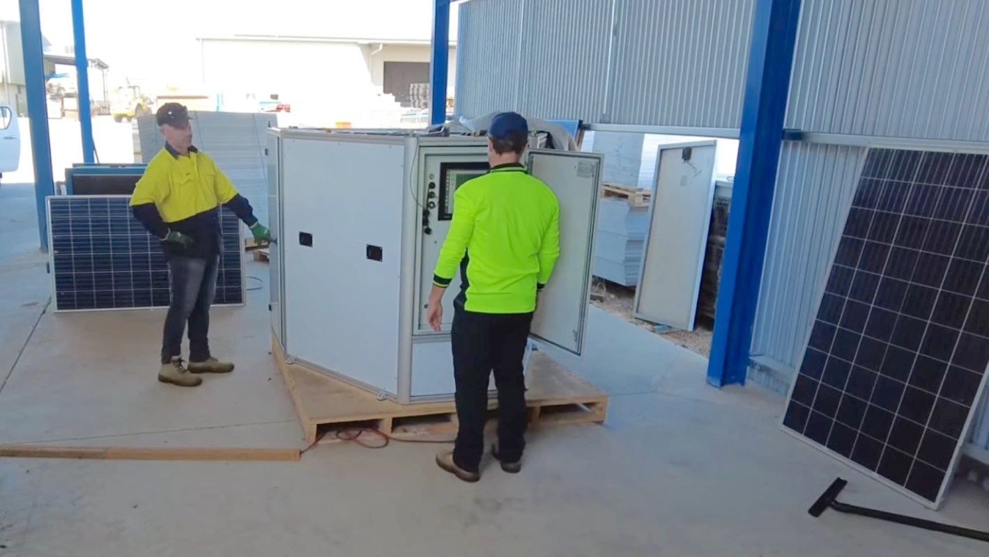 Two workers in safety gear are positioning a white electrical cabinet near solar panels in a storage area.