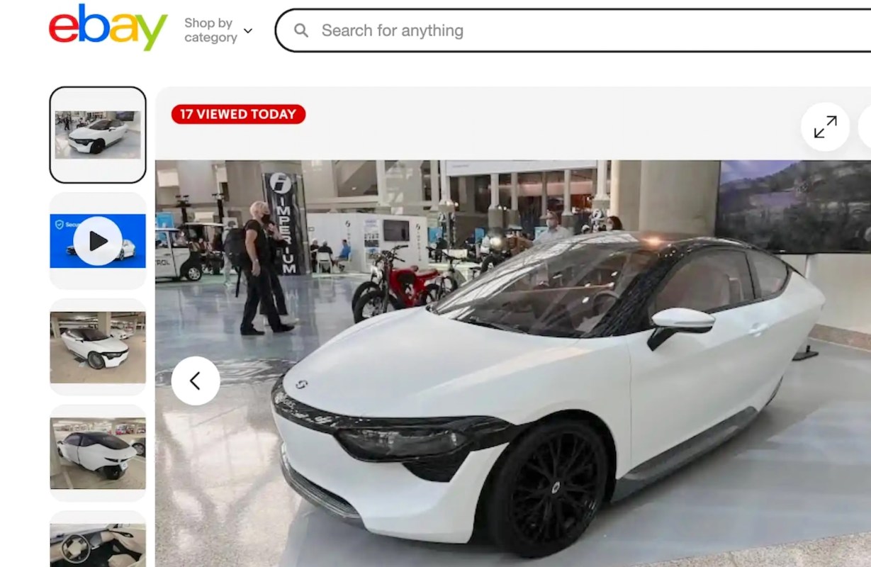 A screenshot of an eBay listing of a white electric vehicle in an exhibition hall with other vehicles in the background.