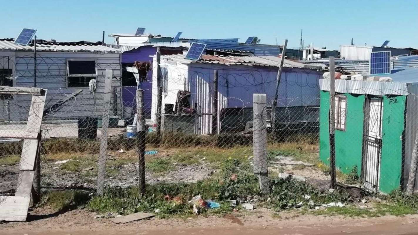 A series of makeshift homes surrounded by a fence, with solar panels on rooftops and litter in the foreground.