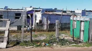 A series of makeshift homes surrounded by a fence, with solar panels on rooftops and litter in the foreground.