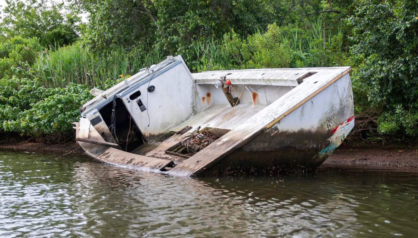 An abandoned boat is partially submerged in a waterway surrounded by lush vegetation.