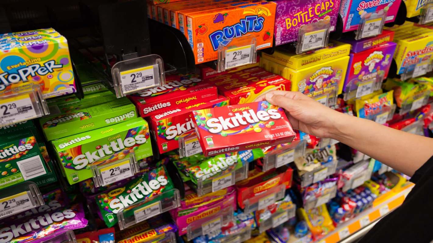 A hand holds a box of Skittles in a display of colorful candy packages.