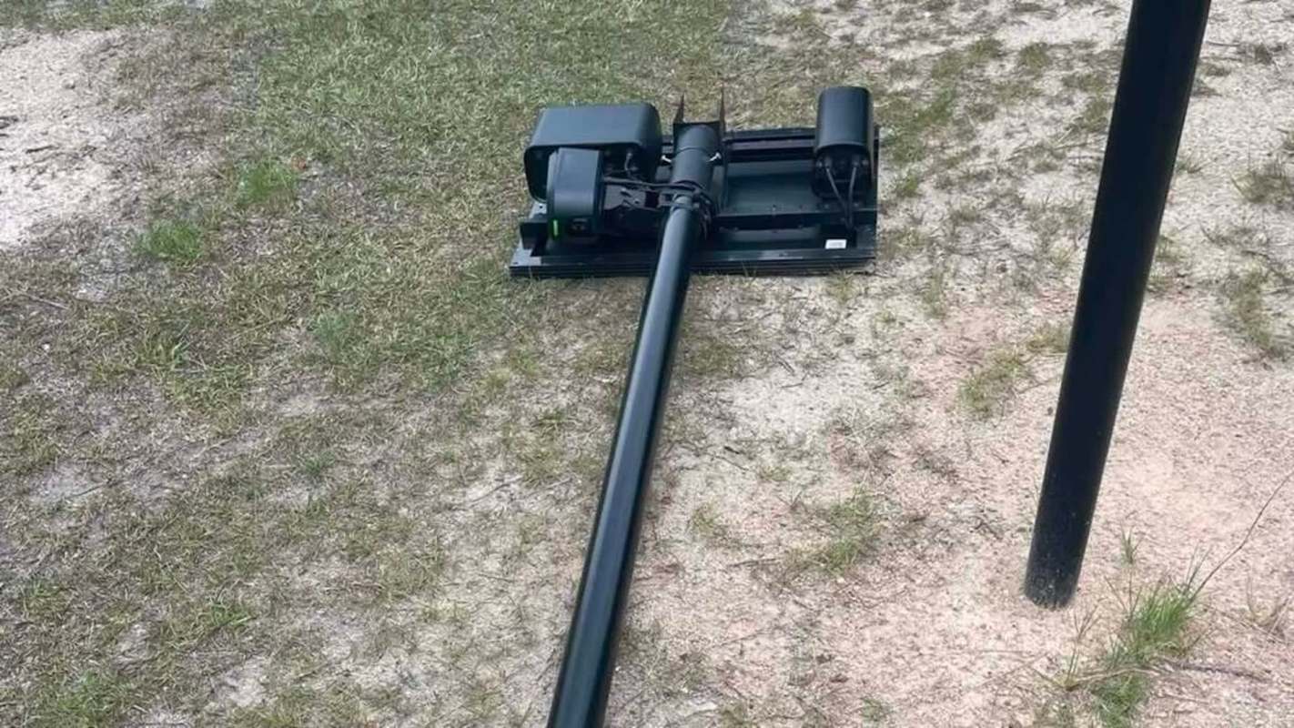 A black Flock camera with a long tube on grass and dirt, next to a vertical pole.