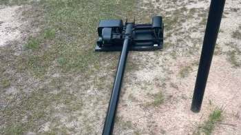 A black Flock camera with a long tube on grass and dirt, next to a vertical pole.