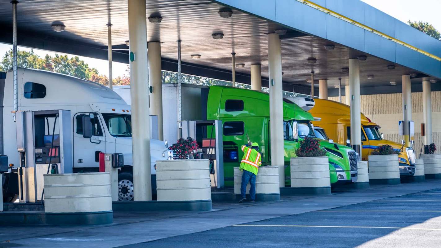 Several trucks are parked at a fuel station, with a person in a safety vest assisting nearby.