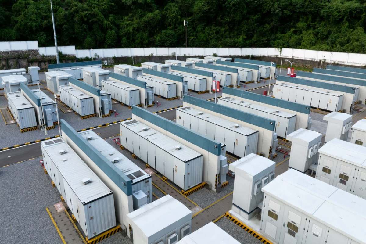 A series of rectangular battery storage units arranged in rows within a gravel area surrounded by green foliage.
