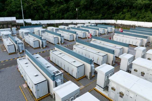 A series of rectangular battery storage units arranged in rows within a gravel area surrounded by green foliage.