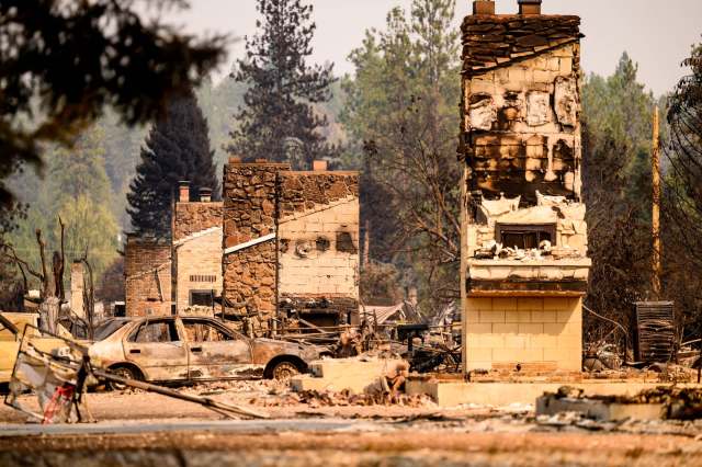 Charred remains of houses and a burned-out car in a devastated area after a wildfire.