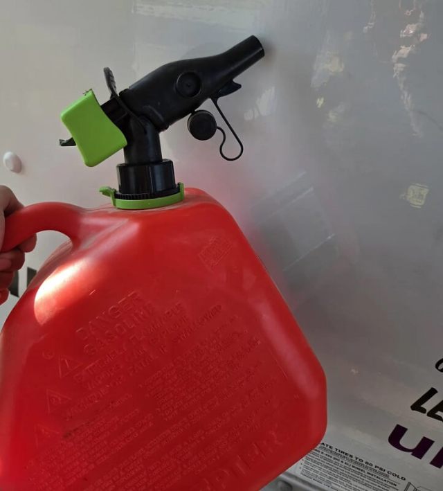 A hand holds a red gas can with a black spout and green nozzle against a light background.