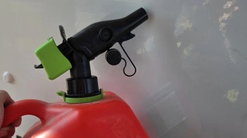 A hand holds a red gas can with a black spout and green nozzle against a light background.