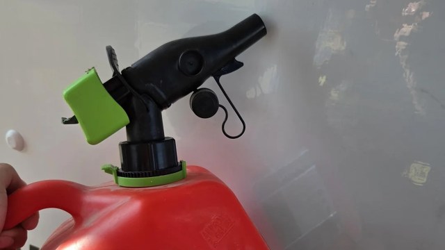 A hand holds a red gas can with a black spout and green nozzle against a light background.