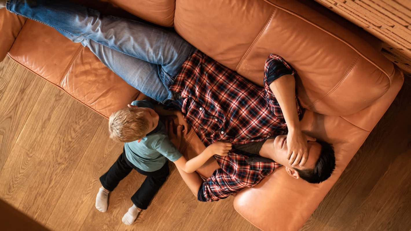A man lies on a couch with his hand over his head while a child plays nearby.