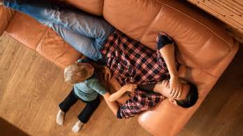 A man lies on a couch with his hand over his head while a child plays nearby.