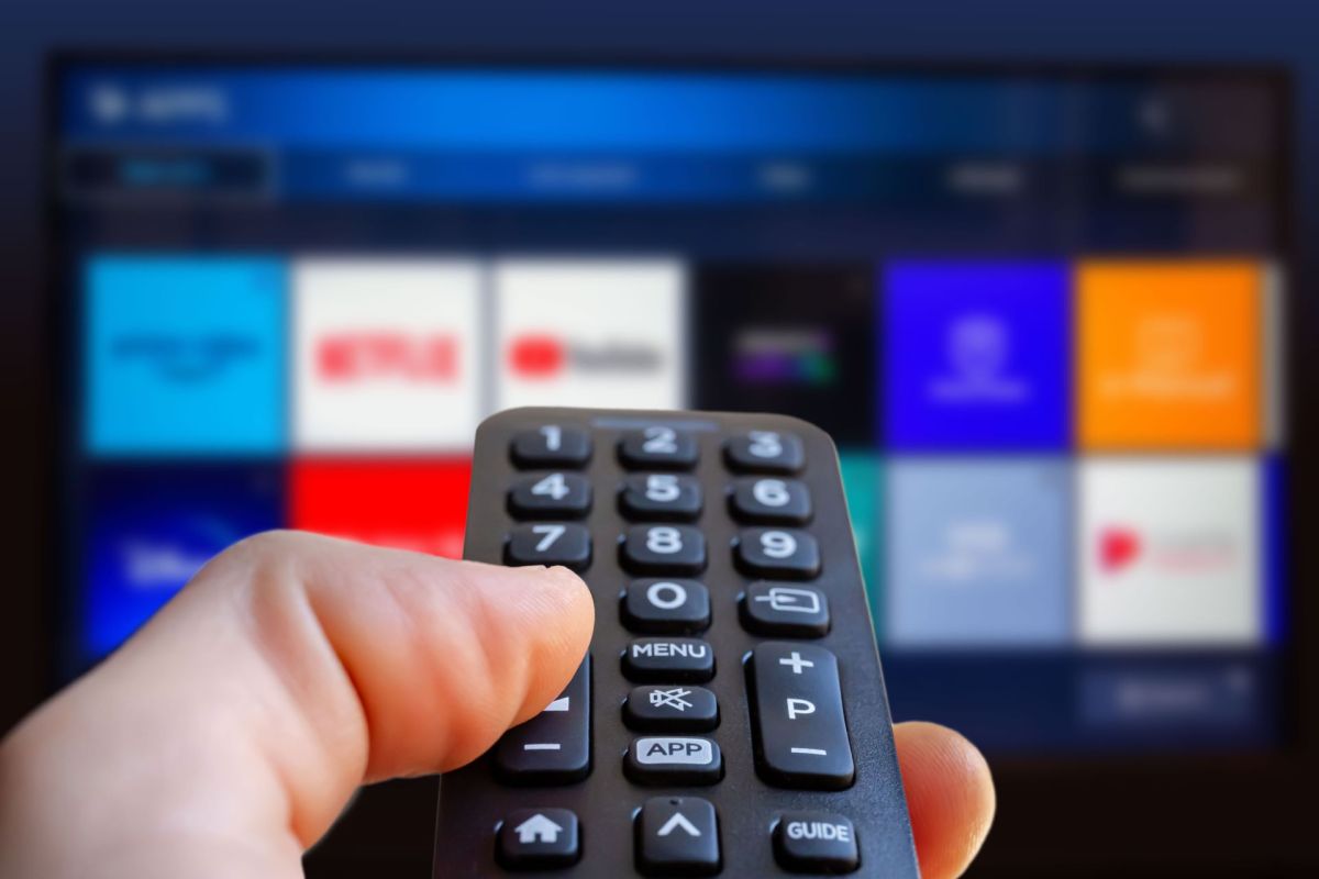 A hand holds a remote control in front of a television displaying various streaming app icons.