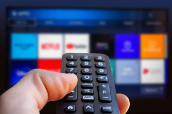 A hand holds a remote control in front of a television displaying various streaming app icons.