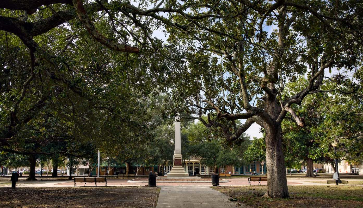A park featuring large trees and a monument surrounded by benches and a pathway.