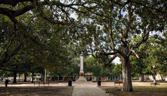 A park featuring large trees and a monument surrounded by benches and a pathway.