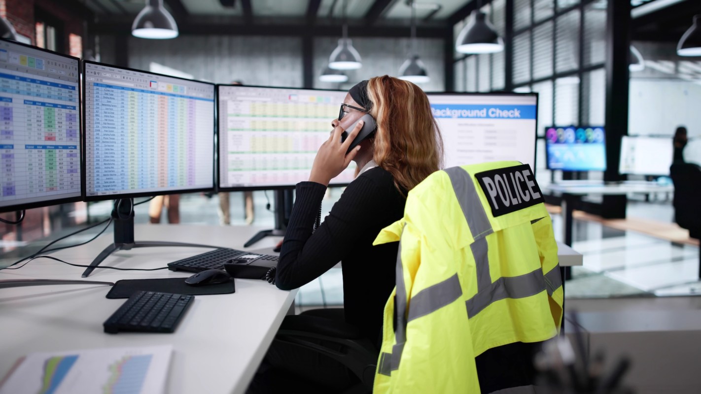 A person speaks on the phone while working at a desk with multiple computer monitors displaying data.