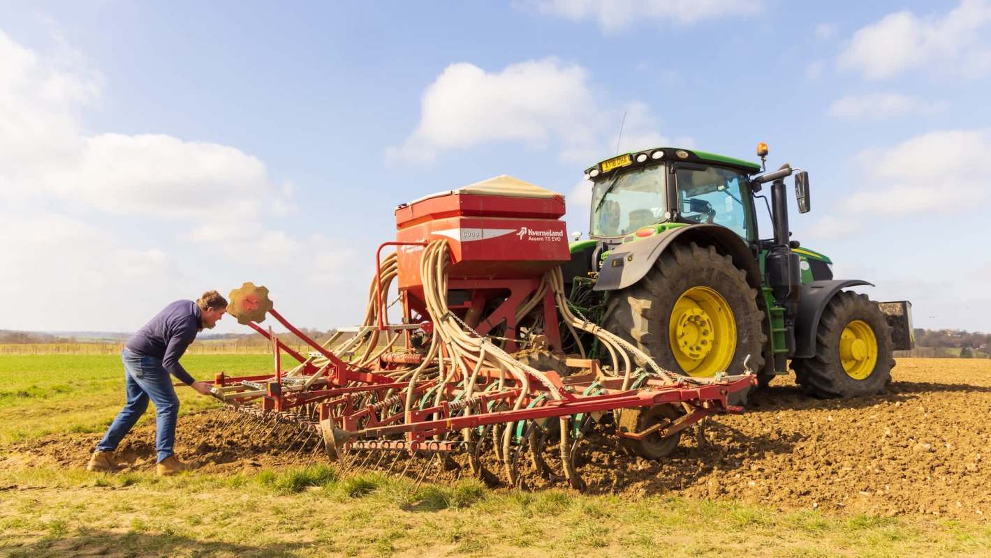 A man operates a seed drill attached to a tractor in a field.