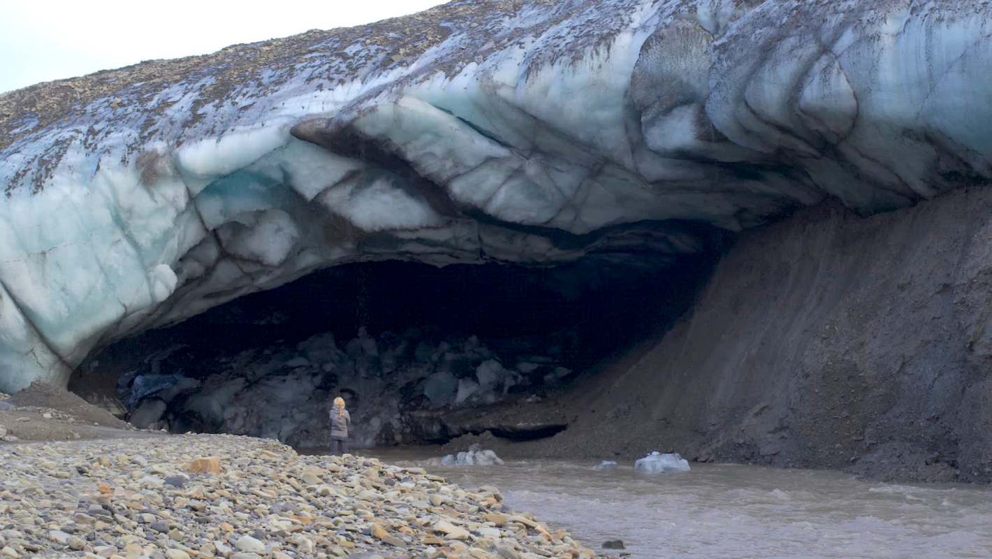 A person standing outside an ice cave near a riverbank with rocky terrain.