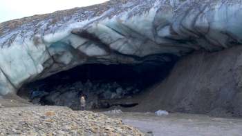 A person standing outside an ice cave near a riverbank with rocky terrain.